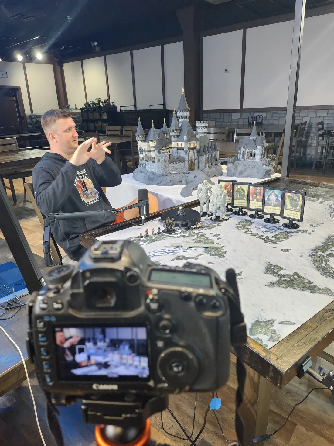 A camera on a tripod in the foreground filming a man at a game table covered in a snowy map and model castles