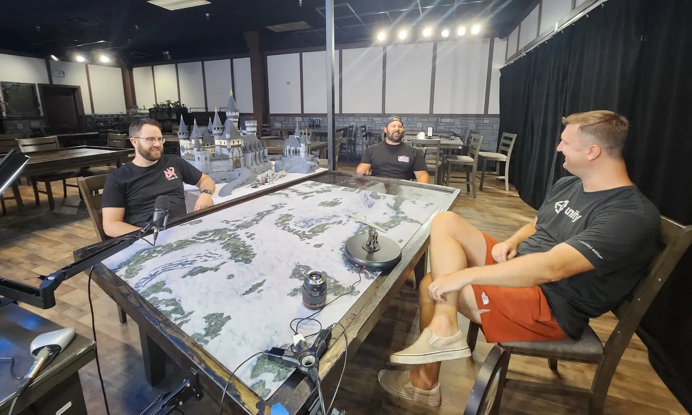 Three people laughing around a game table with a snowy map and model castles, microphones set up for filming