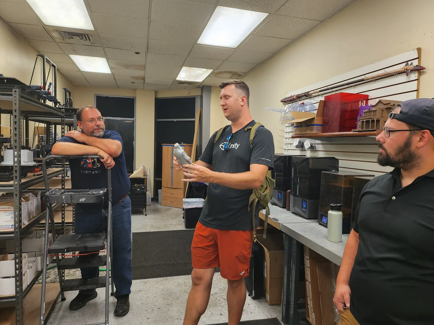 Three people talking in a room lined with 3D printers, one holding a freshly printed miniature