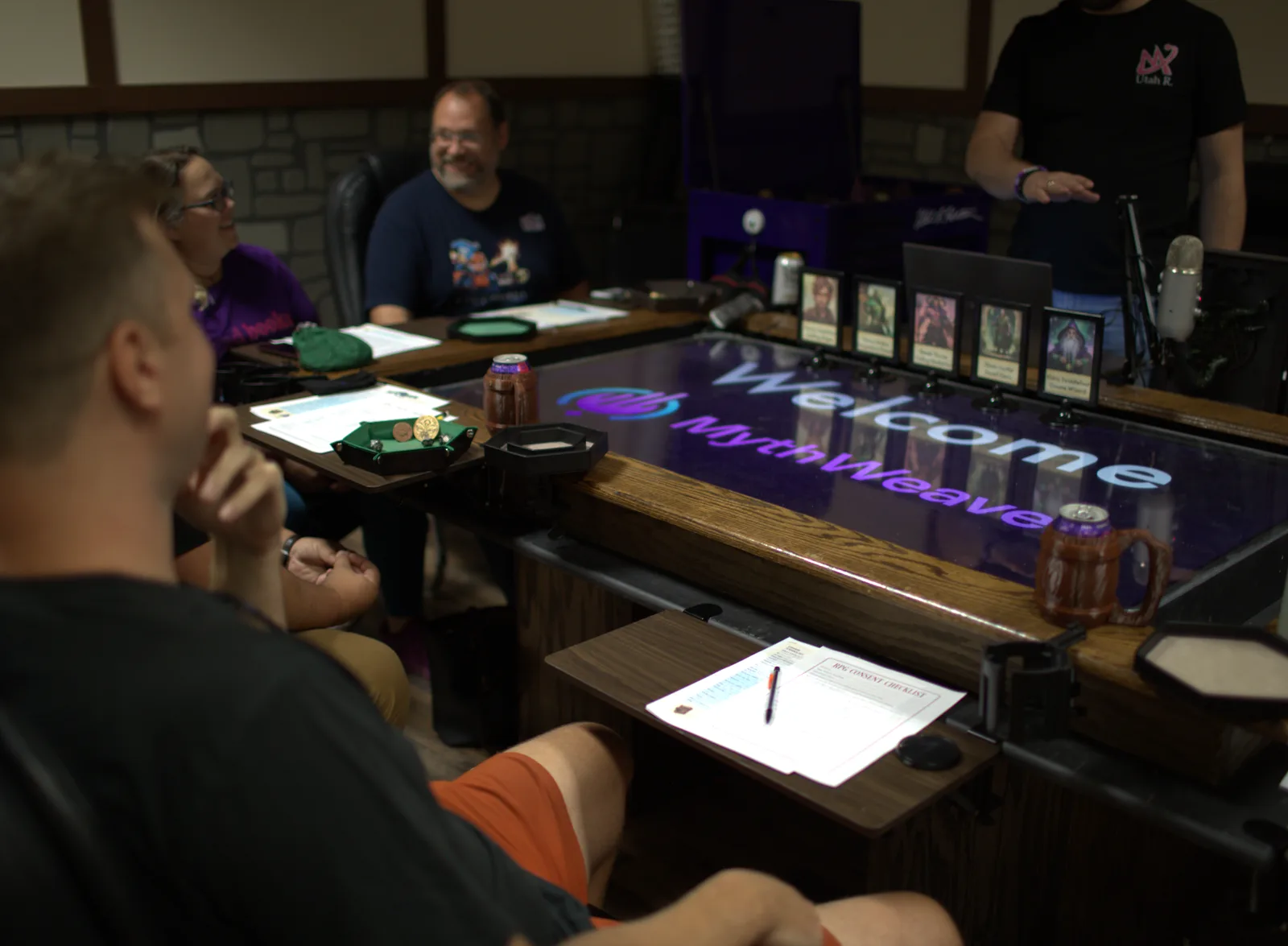 Four people around a large digital gaming table lit with the MythWeaver welcome screen